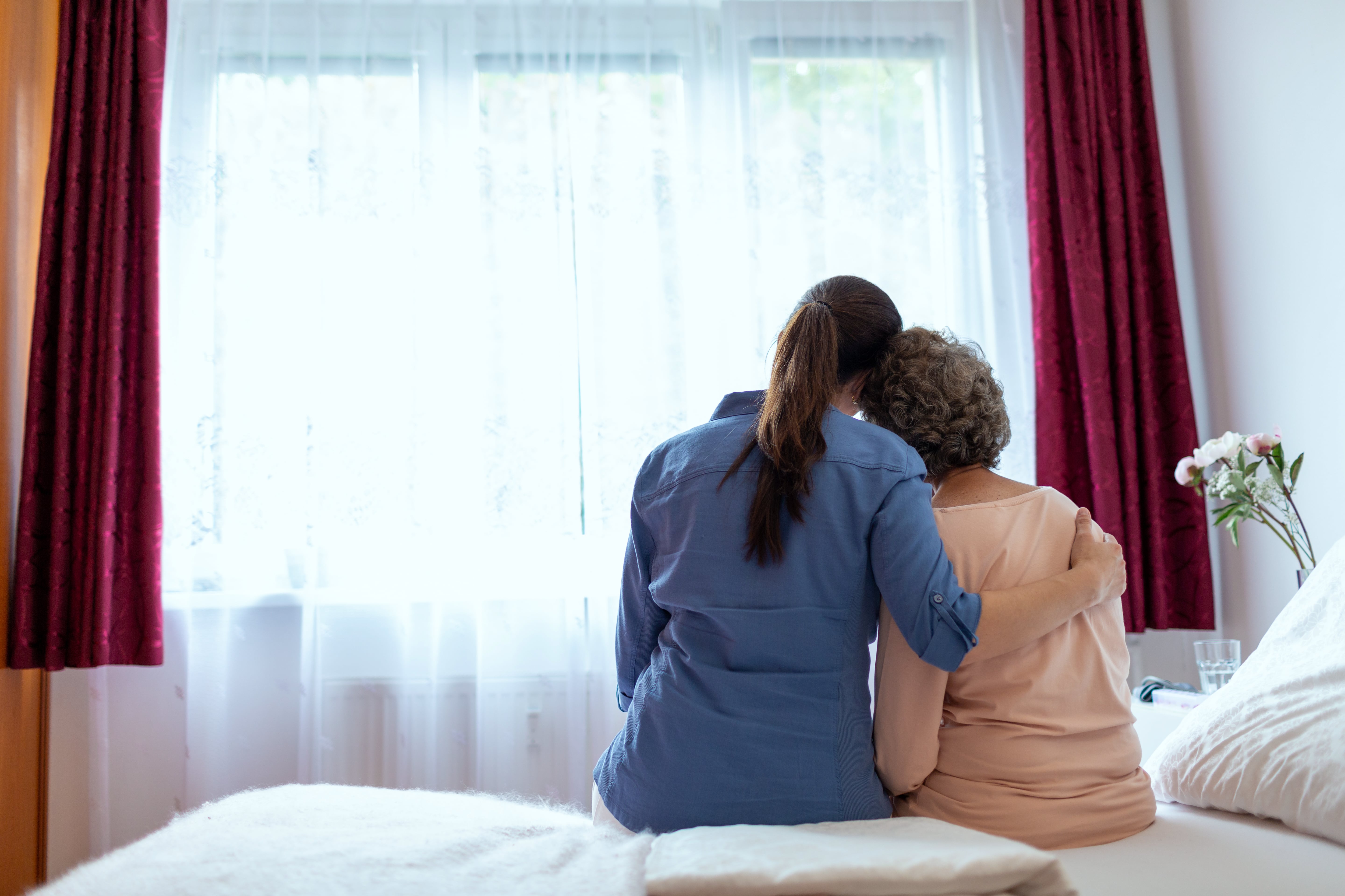 A back view of a caregiver and older loved one embracing while sitting on a bed
