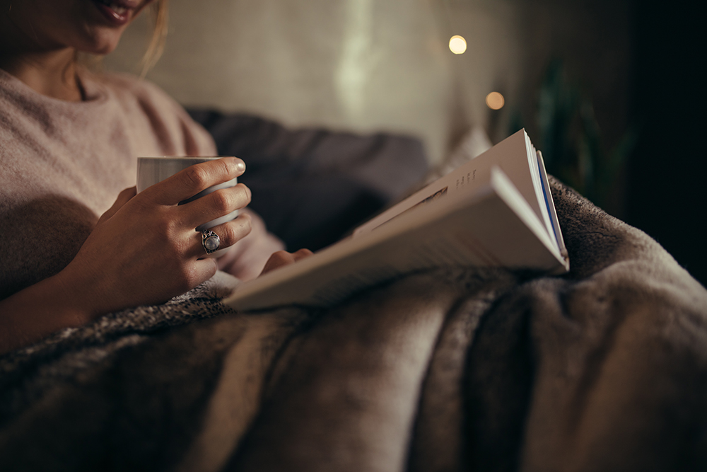 A caregiver relaxing under a blanket with a book and cup of tea